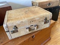 Overall front and top view of vintage beige hard-shell suitcase sitting on wooden surface with other vintage suitcases visible behind. Shows general condition and form of the suitcase.
