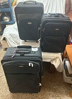 Three black soft-sided suitcases arranged in a triangular layout on carpet and a table, showing front sides with zippers and handles visible.