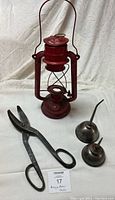 Photo shows a red vintage kerosene hurricane lamp, two small metal thumb oiler cans, and a large forged steel pair of tin snips on a white background.