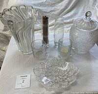 Photo showing the full set of vintage crystal and glass items on white cloth, including large wavy vase, tall vase, two mugs, lidded container with floral etching, textured dish and brown cylindrical item.