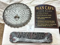 Set of three metal man cave signs on white fabric background. Includes bottle cap shaped welcome sign with bottle opener, man cave rules sign, and wrench design sign.