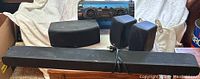 Photo showing the set of five speakers including soundbar, curved home theater speaker, two small cube speakers, and blue boombox speaker arranged on a wooden table.
