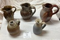 Five vintage ceramic pitchers of different sizes, colors ranging from brown to gray, placed on a white cloth surface.
