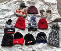 Full lot photo showing 11 winter hats/beanies and one pair of gloves laid out on crumpled white paper.