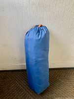 Blue drawstring carry bag containing the 4 person tent, standing upright against a wall.