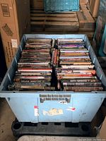 Blue industrial tote filled with DVDs arranged in rows; titles include 'City of Ghosts,' 'Fracture,' and others visible on the spines.