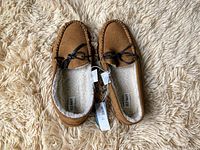 Top view of brown moccasin-style slippers with fleece lining and black bow detail, tags attached indicating new condition.