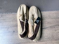 Top and side view of men's moccasin style slippers in beige with brown trim and fleece lining, showing brand tag attached on right slipper.