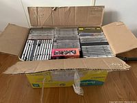 Overhead photo showing open cardboard box tightly packed with multiple rows of music CDs in jewel cases, some with visible spines of different colors.