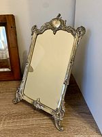 Vintage rectangular mirror with ornate silver-colored metal frame and stand, placed on wooden surface near corner wall.