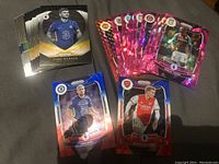 First image shows a stack of Panini Prizm soccer cards including several shiny red, pink, and purple parallel cards with rosters of players from top European teams. Some key players and rookies featured.