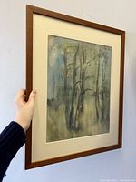 Photo showing framed painting being held, displaying impressionist trees in subdued colors.