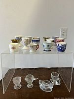 Porcelain and ceramic egg cups displayed on clear acrylic riser, showing a variety of colors, patterns, and gold detailing.