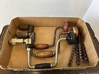 Five antique hand drills with wood handles and twisted auger bits laying in a cardboard box.