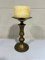 Vintage large brass candle holder with pillar candle, placed on a gray surface with a wall background.