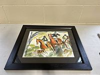 Framed abstract equestrian art print featuring three riders and a dog in muted tones, shown on a table against a plain wall.