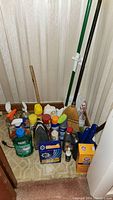 Overview of cleaning supplies and household tools including iron, spray bottles, broom, mop, and plunger arranged on floor in corner.