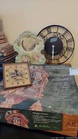 Lot includes three items: a round metal wall clock with Roman numerals and loose hands, a square wind-up clock with beige face and black numbers, and a boxed Medieval Clock building kit with ceramic holder.