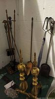 Photo showing full fireplace tool set including brass andirons, shovels, brooms, and pokers against a basement wall.