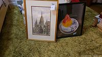 Two framed artworks leaning against a wall on carpet, one depicting Barcelona Cathedral and the other a pear oil painting.