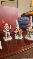 Three gnome figurines from Goebel, showing front and side views on a wooden surface, with a sofa in background.
