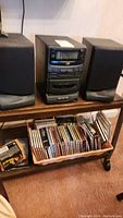Front view of the JVC stereo system with two speakers and a lower shelf filled with assorted CDs in cases and a CD case box on the carpet.