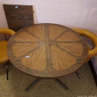 Round brown wood grain patterned laminate table top with petal design, leaf stored behind table. Two mid-century style orange-brown chairs partially visible at sides.