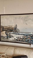 Framed seaside canvas art showing the coastal building and shoreline with waves, signed by Lee Reynolds.