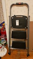 Front view of folded Rubbermaid 3 step ladder showing black textured steps and gray frame with Rubbermaid logo.
