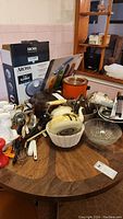 Photo shows assortment of kitchenware items on wooden table including Aroma 3-in-1 grillet box, orange Crock Pot, glass pot, metal utensils, and baking pans.