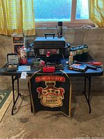 Overview of entire camping lot on black folding table showing portable BBQ, Coleman lantern box, propane canisters lined up, flashlight, lighter, frying pan, spray can, vintage red gas stove, and Smirnoff apron hanging on table front.