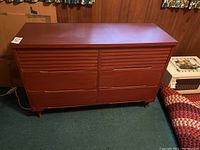 Wood dresser with six drawers closed, reddish-brown finish, four round tapered legs with brass foot caps, shown against wood panel wall and green carpet.