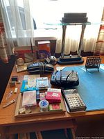 Group of various office supplies arranged on wooden desk near window, including a desk lamp, calculator, stapler, and several small boxes of push pins and labels.