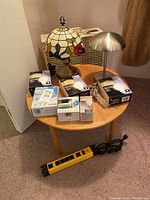 Box fan behind small round wooden table with 2 lamps, multiple boxes of lightbulbs, and a yellow power strip on carpeted floor