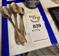 Four silver spoons laid on a white and blue marble surface with a tag showing 91.97 grams weight