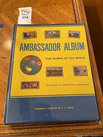 Front cover of the Ambassador Album for stamps of the world showing the yellow and blue hard cover with illustrative stamps and globe graphic.