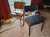 Two Mid-Century Modern chairs in a living room; one with teak wood frame and blue fabric upholstery, the other with brass frame and cracked brown vinyl upholstery.
