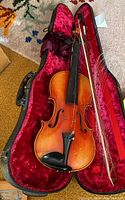 Violin inside black case lined with red velvet along with a bow and loose violin strings