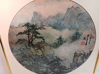 Close-up of circular watercolor painting with mountain landscape, trees, mist, and artist signature/stamp in Asian characters.