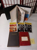 Display of items includes three Playbills with one from In the Heights and two Grease editions, an antique hymnal, a red book labeled Vital Fault, and an album standing upright with visible pages.
