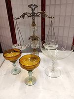 Photo showing the crystal and brass scales of justice centerpiece with one pendant missing, two amber art glass goblets, and the clear stemmed crystal bowl on a white surface in front of a red folding screen.