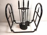 Set of fireplace tools with black handles arranged on a circular stand, placed inside black metal log holder with spear-like decorative finials on each side.