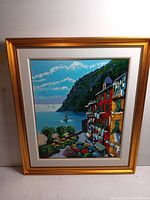 Full view of framed serigraph print depicting seaside village with gold-tone frame and white mat