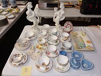 Wide view showing ceramic cherub candle holders, assorted fine china teacups and saucers, artisan seashell tray, and Wedgwood jasperware boxes all arranged on table