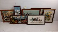 Overview of all seven framed needlepoint embroidery pieces, showing varied landscape and floral scenes, with different frames mostly in wood tones.