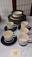 Photo showing stacked plates, bowl on top, and five cream and dark blue mugs with matching saucers in front.