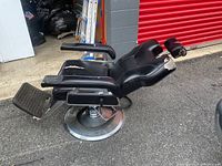 Side view of the black reclining barber chair showing heavy metal circular base and black armrests with visible upholstery damage on seat and headrest.