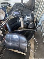Six black barber chairs stacked and arranged showing padded vinyl seats, metal frames, chrome hydraulic bases, footrests, and black armrests. Condition shows scratches and seat creases.