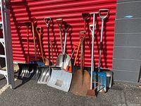 Photo showing 14 garden tools, including various shovels, hoes, and cultivators, leaning against a red building wall.