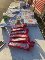 Overview photo of assorted boxed kids learning activity kits, paint palette, clear containers with beads and toys, and 7 red mesh pouches with milkshake straws arranged on a table outside.